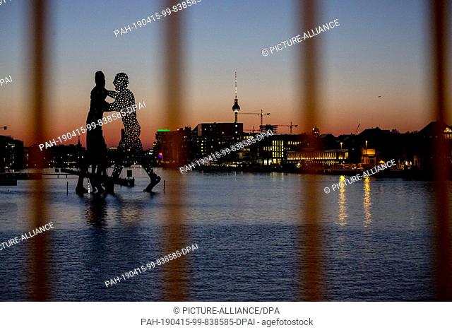 15 April 2019, Berlin: The statue Molecule Man and the Berlin television tower can be seen at dusk through the railing of the Elsen Bridge