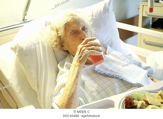 Elderly woman lying in a sickbed, drinking from a mug, in front of her a plate of food