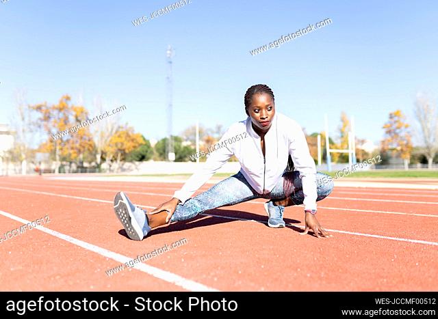 Young sportswoman looking away while doing warm up exercise on sports track against clear sky