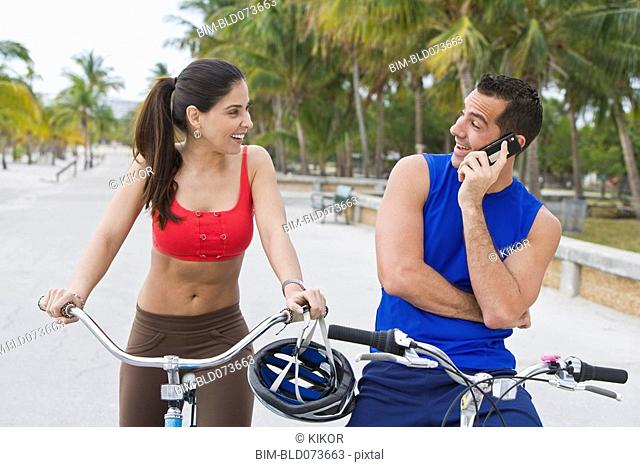 Hispanic man on bicycle talking on cell phone while girlfriend waits