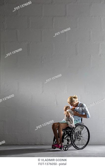 Full length of woman breastfeeding baby girl in wheelchair against gray wall