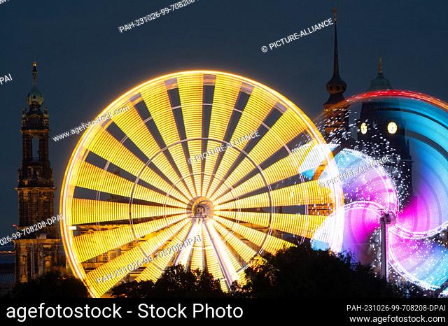 25 October 2023, Saxony, Dresden: A Ferris wheel and and the Booster Maxx carousel are illuminated with colorful lamps in the evening at the autumn festival on...
