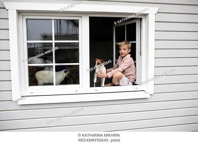 Portrait of boy sitting on window sill with Jack Russel Terrier and Siam cat looking out of open window
