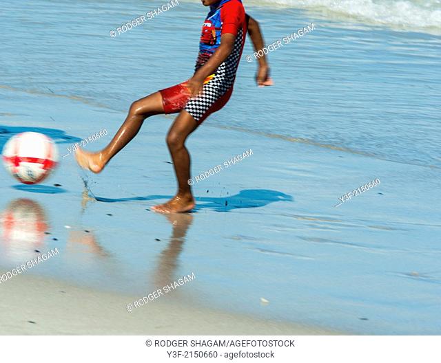 A young African lad dressed in a sun-protective bathing suit, kicks a ball on the beach at low tide. Cape Town South Africa