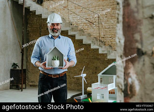 Happy architect with house model and potted plant standing at construction site