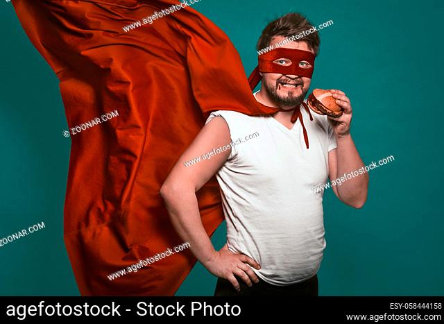 Hungry super hero man eating a hamburger, super hero or antihero in a red suit with a flying cloak eats fast food looking at the camera