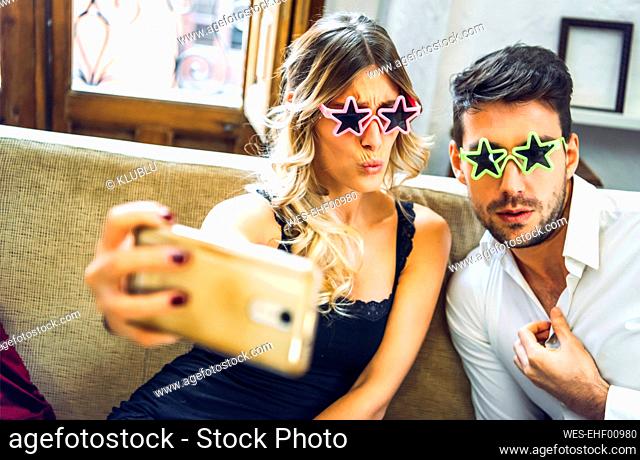 Playful young couple wearing comedy glasses and taking selfie on couch in living room at home