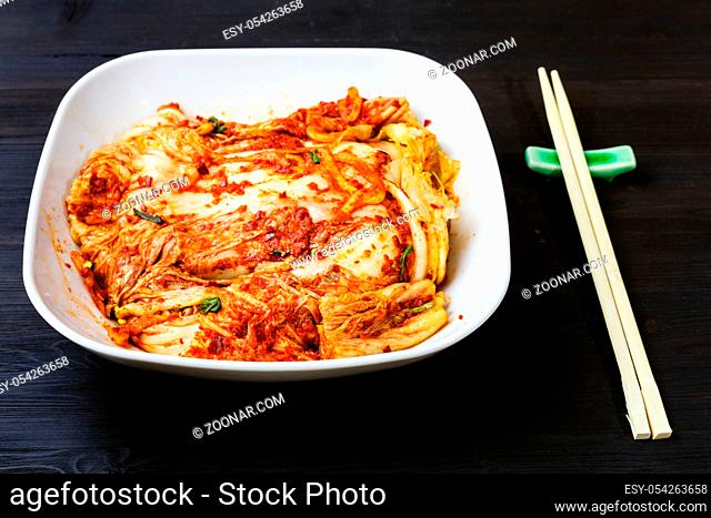 korean cuisine - kimchi appetizer (spicy nappa cabbage) in white bowl and chopsticks on dark brown wooden table
