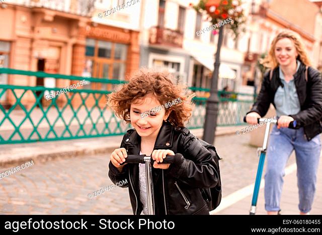 The beautiful little girl with curly hair rides a scooter in the city. Behind her is mom on a scooter as well. Focus on the girl