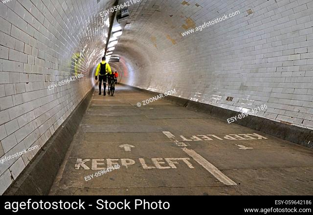Cyclist wearing yellow jacket walking with his bicycle inside Greenwich foot tunnel under river Thames, more blurred pedestrians in background