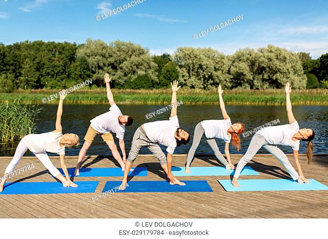 fitness, sport, and healthy lifestyle concept - group of people making left triangle pose on mat outdoors on river or lake berth