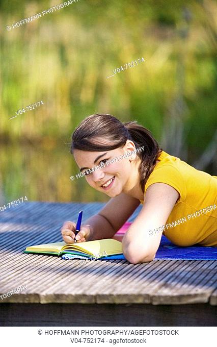 Teenaged girl lying on a footbridge and writing in her diary, smiling at the camera
