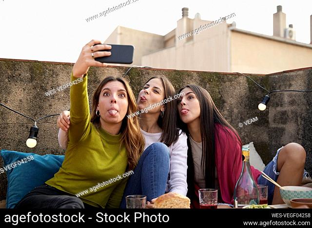 Female friends sticking out tongue while taking selfie on rooftop