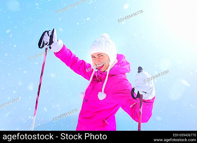 Happy young woman in pink ski jacket holding ski poles in gloves, smiling with sun and snow in background