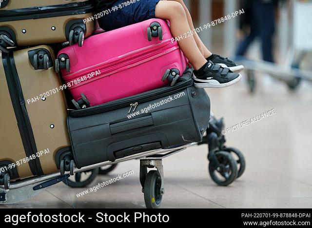 01 July 2022, Hamburg: A child sits on suitcases on a baggage cart in a queue in front of a check-in counter in Terminal 1 at Hamburg Airport