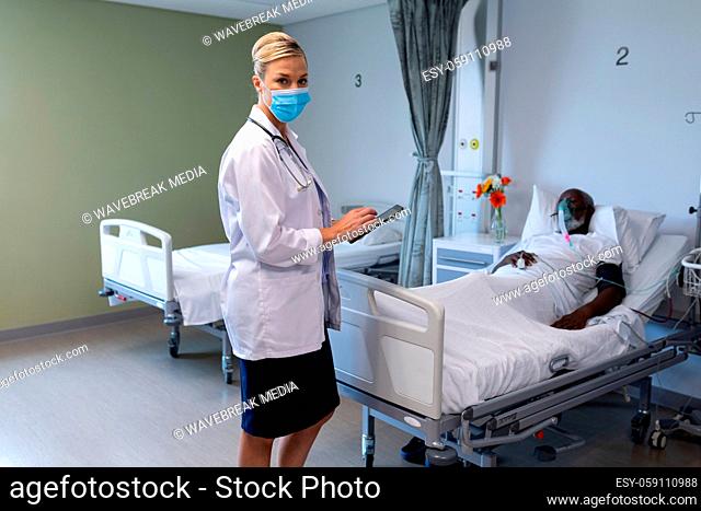 Caucasian female doctor wearing face mask holding tablet looking to camera in hospital patient room