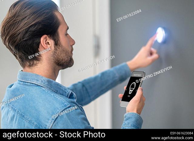 Adjustment. Young bearded man standing sideways to camera looking attentively touching button on wall indoors