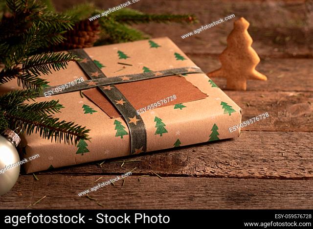 Gift box wrapped in brown classic paper and Christmas trees design, fir twigs, Xmas ball, and a Christmas tree cookie, on a rustic table