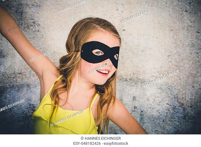 beautiful, blond girl with black superhero mask and yellow shirt is posing in front of concrete background