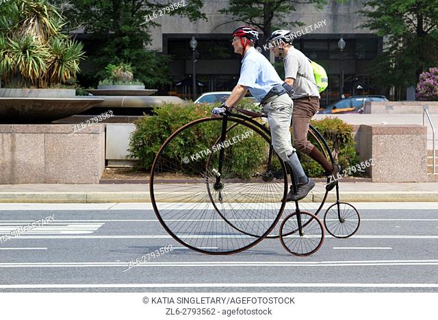 Two caucasian male ride old fashioned bicycles with very big wheels and small wheels. They are riding penny-farthing' bike