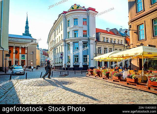 TALLINN, ESTONIA - JULY 14, 2019: Tourists sitting in street restaurant in old town of Tallinn, statue of walking man and cityscape in evening sunshine