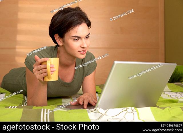 Woman lying on bed using laptop computer holding coffee mug