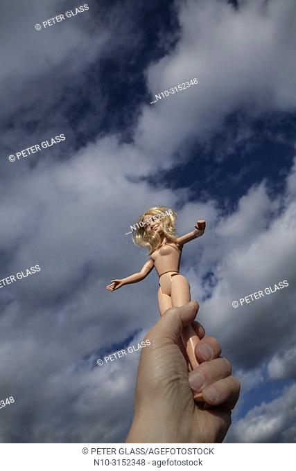 Man's hand holding a naked blonde female doll against a cloudy sky