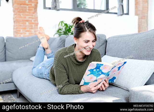 Smiling woman reading book while lying on sofa at home