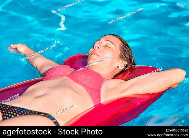 Portrait of pretty young woman in swimsuit lying on red inflatable mattres at swimming pool