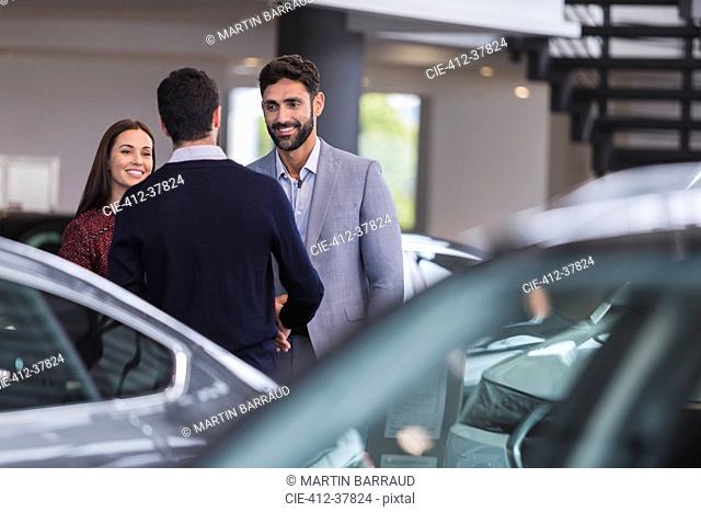 Car salesman greeting, shaking hands with couple customers in car dealership showroom