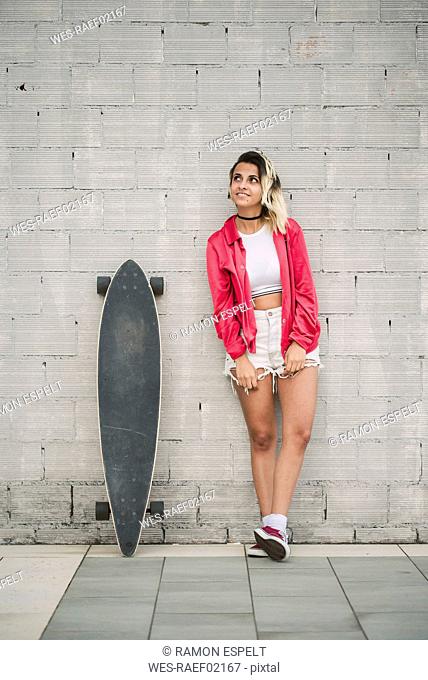 Portrait of a young woman with her long board, in front of a wall