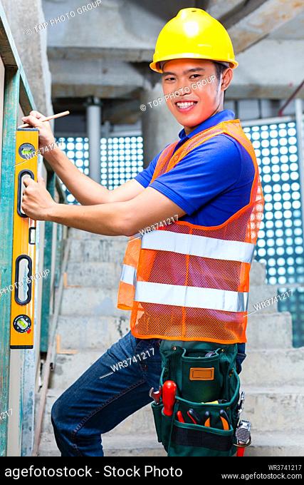 Asian Indonesian builder or craftsman with hardhat and bubble level controlling or checking a wall of a tower building or construction site