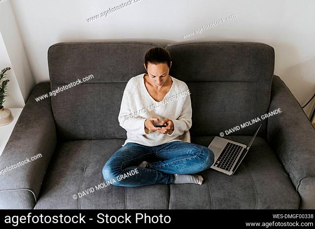 Woman using mobile phone while sitting with laptop on sofa