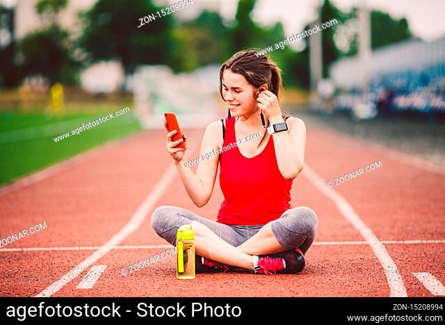 theme healthy lifestyle, sports music. beautiful young Caucasian woman rests sitting treadmill running rubber stadium uses technodogy mobile phone hand insert...