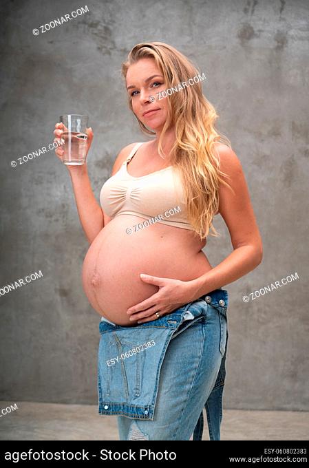 Pretty pregnant woman in denim jumpsuit and bra holding glass of water while standing over gray wall background. Maternity, pregnancy
