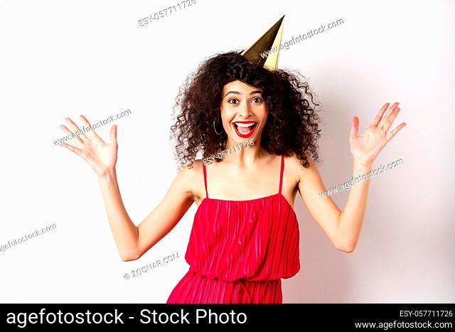 Cheerful young woman in red dress, celebrating birthday, wearing party hat and smiling, screaming of joy, standing on white background