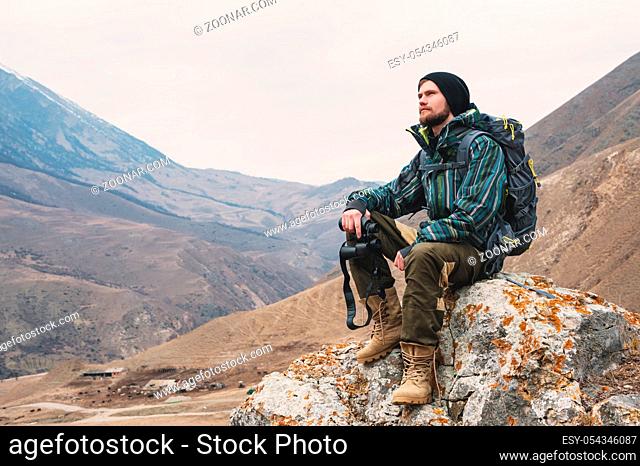 Tired bearded hipster with binoculars in his hands sits on a stone among the Caucasian mountains and looks out into the distance