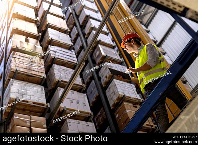 Worker wearing hardhat checking inventory standing with tablet PC by railing in warehouse