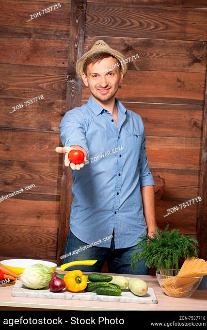 Chef holding tomato in his hand on wooden. Smiling man with straw hat on preparing food with fresh vegetables