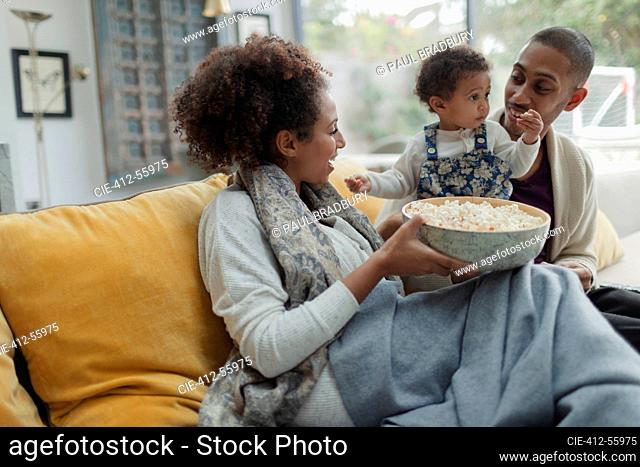 Happy parents and baby daughter watching TV with popcorn on sofa