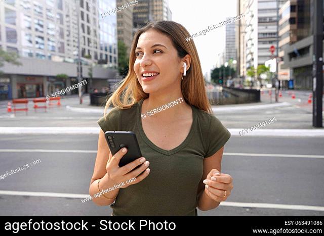 Cute Brazilian Caucasian girl with earphones wireless and mobile phone looks to side in modern metropolis. City life concept