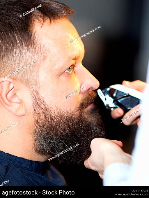 Man having his mustache and beard trimmed at a barber shop, close up side view of his head and the clippers or razor