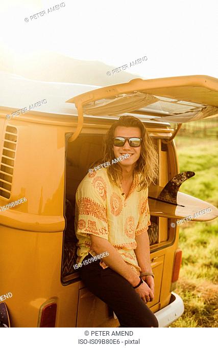 Portrait of young male surfer with long hair leaning against vintage recreational vehicle, Exeter, California, USA