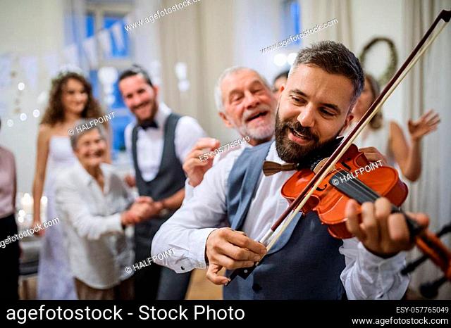 A mature man playing a violin on a wedding reception, bride, groom and other guests dancing