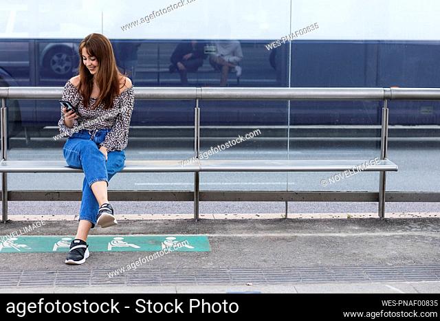 Smiling young woman using smart phone while sitting on bench at tram station