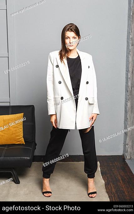 Young elegant woman in white oversize blazer and black ripped pants. Interior portrait