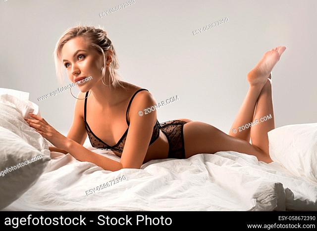 Romantic sensual blond female in black lace bra and panties lying on bed looking away on gray background