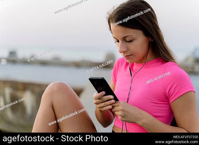 Athlete with in-ear headphones using mobile phone while sitting on retaining wall against sea