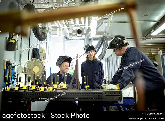 Two students and a lecturer wearing overalls and welding masks at a workbench in a workshop