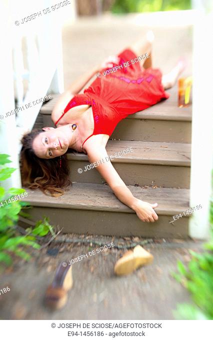 A 36 year old brunette woman in a red summer dress lying on her back on porch steps looking directly at camera